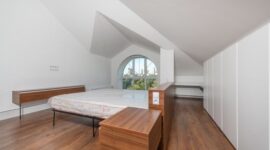 Sebuah kamar tidur minimalis menampilkan tempat tidur rendah dengan seprai putih bersih, meja samping kayu, dan pot tanaman hijau, menciptakan suasana yang tenang dan rapi. Desainnya mengoptimalkan cahaya alami dan ruang penyimpanan tersembunyi untuk fungsionalitas maksimal.