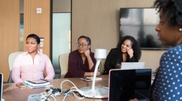 Seorang wanita profesional berdiskusi dengan mentornya di lingkungan kantor modern, menunjukkan kolaborasi dan dukungan dalam pengembangan karier. Gambar ini merepresentasikan pentingnya bimbingan dan pembelajaran berkelanjutan bagi wanita dalam meraih sukses.