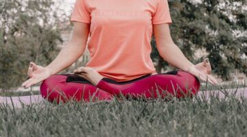 Seorang wanita sedang bermeditasi dengan tenang di tengah hijaunya alam, merefleksikan esensi gaya hidup "slow living" yang menenangkan jiwa dan pikiran dari hiruk pikuk kehidupan. Pemandangan ini menggambarkan ketenangan serta keseimbangan yang bisa dicapai melalui kesadaran penuh terhadap diri sendiri dan lingkungan sekitar.